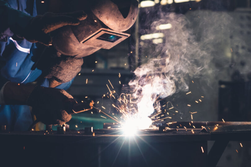 Skilful metal worker working with arc welding machine in factory while wearing safety equipment