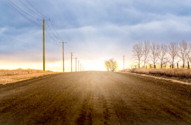 Sunrise scene with sun rays over a county gravel road as wooden power poles and power lines stand along a ditch with natural prairie grasses in Rocky View County Alberta Canada.
