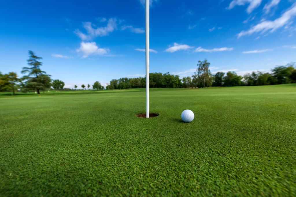 White ball on green grass close to hole with flag against blue sky and green trees