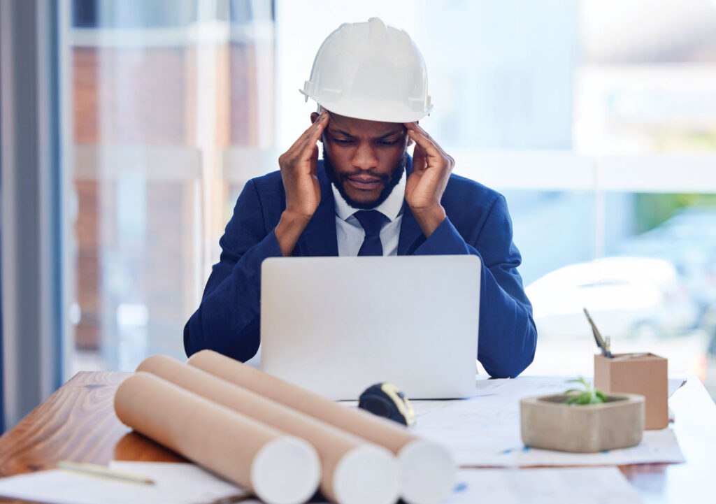 Man sitting at desk wearing white hard hat, holding temples looking at laptop.