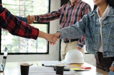 People shaking hands with construction helmet and papers on table between them