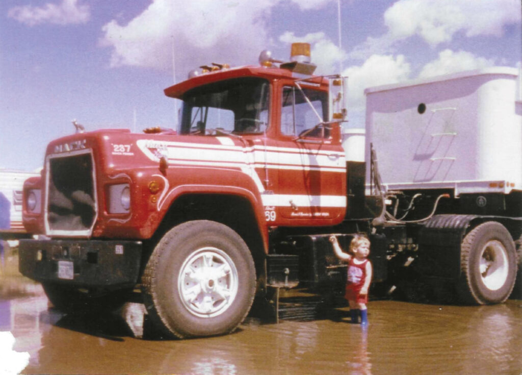 Keegan Urick as a small child in rubber boots standing in water holding on to truck