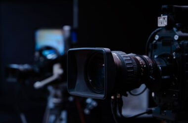 Professional video camera with a lens hood and prime lens focusing on the foreground, with another blurred camera setup in the background