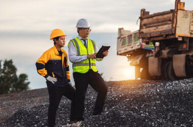 Two construction workers discuss project details on a gravel site during sunset.