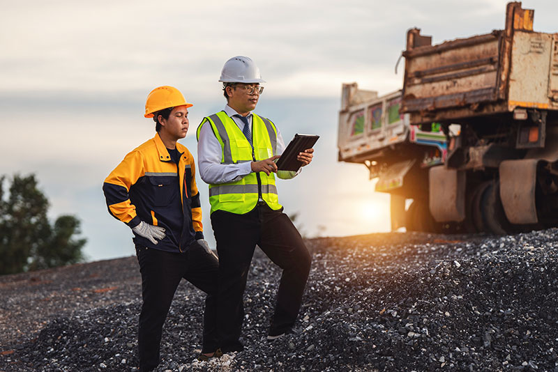 Two construction workers discuss project details on a gravel site during sunset.