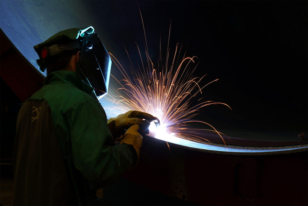 Person welding very large steel pipe with blue tones and orange sparks with nice leading pipe line