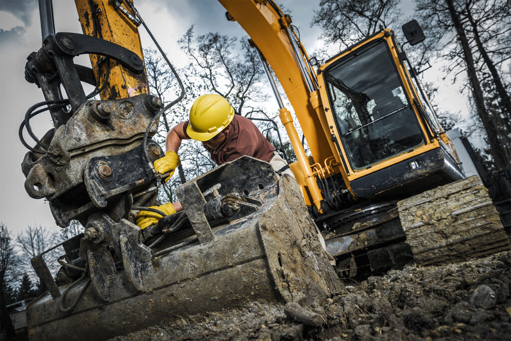 An excavator operator in a hard hat and gloves adjusts machinery in muddy conditions at a construction site surrounded by trees during daylight hours
