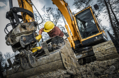 An excavator operator in a hard hat and gloves adjusts machinery in muddy conditions at a construction site surrounded by trees during daylight hours